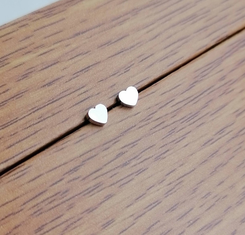 May include: A pair of silver heart-shaped stud earrings on a wooden surface.