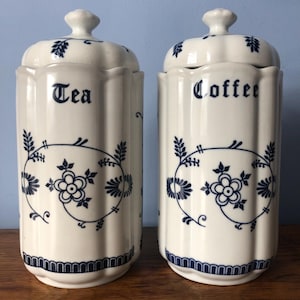 May include: Two off-white ceramic canisters with matching lids, each featuring a vertical fluted design and a dark blue floral vine pattern. One canister is labelled Tea and the other is labelled Coffee in a black serif font. Both have a blue border.