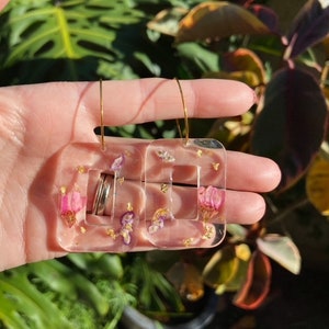 Könnte beinhalten: Ein Paar quadratische Ohrringe mit goldenen Creolen. Die Ohrringe sind aus klarem Harz mit rosa und goldenen Flocken und getrockneten Blumen.
