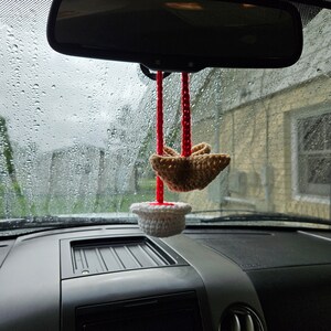 May include: A white crocheted cupcake with red string hanging from the rearview mirror of a car. A brown crocheted bird is hanging from the string above the cupcake.