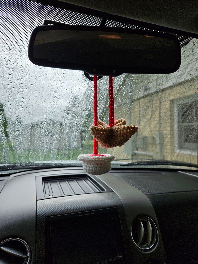 May include: A white crocheted cupcake with red string hanging from the rearview mirror of a car. A brown crocheted bird is hanging from the string above the cupcake.