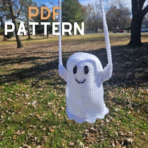 May include: A white crocheted ghost with black eyes and a smiling mouth hangs from a string. The ghost is in front of a blurry background of green grass and brown leaves. The text "PDF PATTERN" is in the upper left corner of the image.