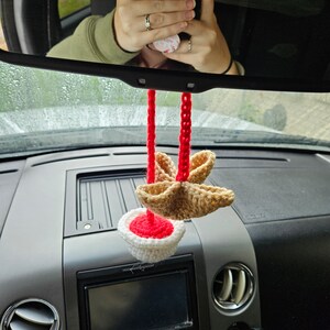 May include: A crocheted car air freshener with a red and white base and a brown star-shaped top. The air freshener is hanging from the rearview mirror of a car.
