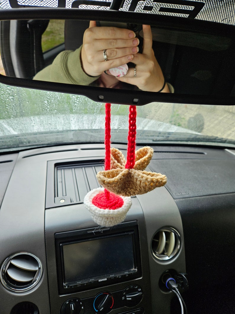 May include: A crocheted car air freshener with a red and white base and a brown star-shaped top. The air freshener is hanging from the rearview mirror of a car.