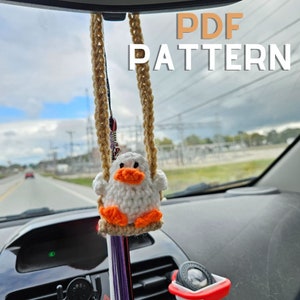 May include: A white crocheted duck with an orange beak and feet is hanging from the rearview mirror of a car. The duck is on a swing made of brown yarn. The text "PDF PATTERN" is visible at the top of the image.