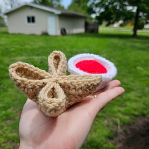 May include: A crocheted amigurumi food set featuring a tan-colored croissant and a white and red-colored strawberry.