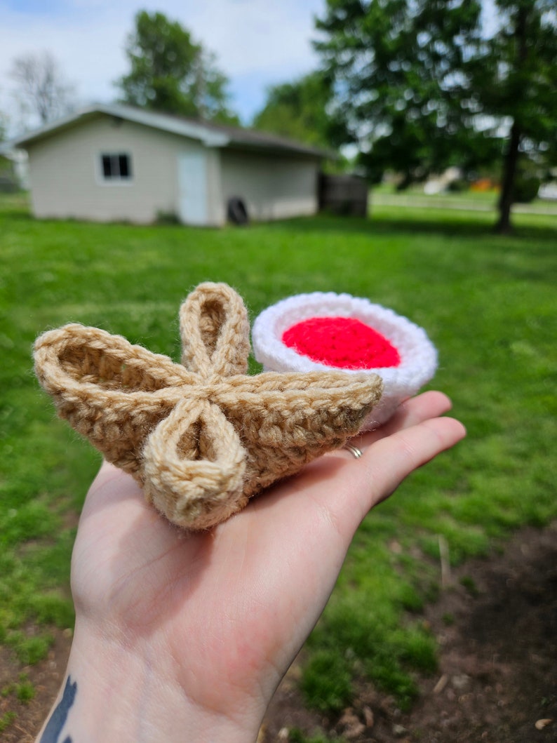 May include: A crocheted amigurumi food set featuring a tan-colored croissant and a white and red-colored strawberry.