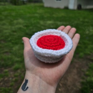 May include: A crocheted red and white pie dish, with a red crocheted filling, is held in a person's hand.