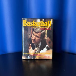 May include: A book titled "Basketball" with a yellow and red title. The cover features a man in a suit, with text that reads "MULTIPLE OFFENSE AND DEFENSE BY DEAN SMITH" and "FOREWORD BY BOB KNIGHT". The book is on a black surface with a blue background.