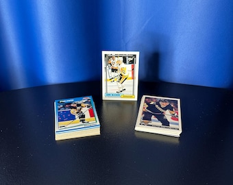 Puede incluir: Tres montones de cromos de hockey con jugadores en acción. Las tarjetas tienen diseños coloridos con logotipos de equipos y nombres de jugadores. El fondo es azul liso.