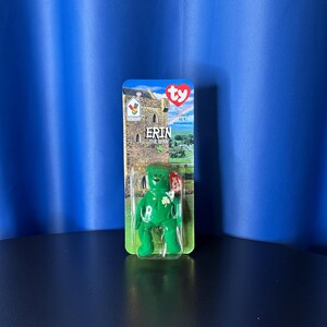 May include: A green Erin the Bear Beanie Baby toy in its original packaging. The packaging features a castle illustration and the text "Erin the Bear". The bear has a shamrock on its chest.