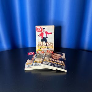 May include: A stack of Elvis Presley "TV Guide" magazines. The magazines feature Elvis on the cover, with the title "Elvis Forever!". The magazines are stacked on a black surface with a blue backdrop.