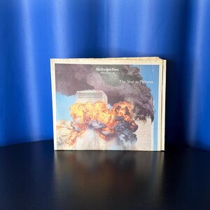 May include: A copy of The New York Times titled The Year in Pictures, featuring a front-page photograph of the World Trade Center towers with orange flames and thick black smoke billowing from the structures against a clear blue sky background.