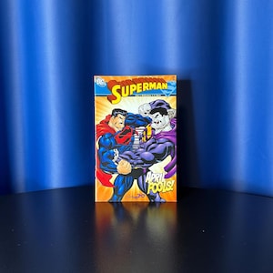 May include: A Superman comic book featuring a vibrant illustration of Superman in a battle scene with another character. The cover has the title "Superman" and the text "April Fools!" in bold letters. The comic is set against a blue backdrop.