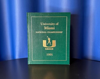 May include: A green book with gold lettering and a gold border. The text reads "University of Miami NATIONAL CHAMPIONSHIP" with a logo and the word "MIAMI" and the year "1991". The book is on a black surface.