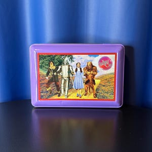 May include: A purple rectangular lunchbox featuring a colorful image of characters from The Wizard of Oz. The image shows Dorothy, the Tin Man, the Scarecrow, and the Cowardly Lion walking on a yellow brick road.