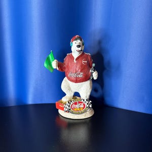 May include: A Coca-Cola polar bear figurine wearing a red racing jacket and hat, holding a green flag and a Coke bottle. The figurine stands on a base with a checkered flag and the Coca-Cola logo.