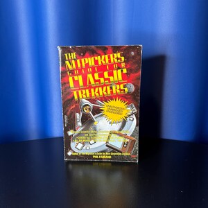 May include: A rectangular book titled "The Nitpickers Guide for Classic Trekkers" with a red, yellow, and black design. The book cover features images of a spaceship and a retro television. The author's name, Phil Farrand, is also visible.