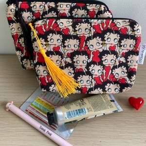 Pu&ograve; includere: Due pochette con cerniera nera con una stampa rossa e nera di personaggi dei cartoni animati. La pochette pi&ugrave; piccola ha un fiocco giallo. Le pochette sono su una superficie di legno con una penna rosa, un tubo di crema per le mani e un cuore rosso.
