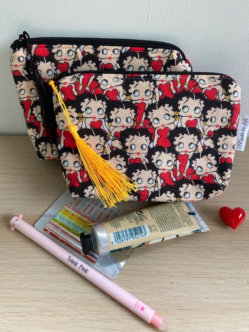 Pu&ograve; includere: Due pochette con cerniera nera con una stampa rossa e nera di personaggi dei cartoni animati. La pochette pi&ugrave; piccola ha un fiocco giallo. Le pochette sono su una superficie di legno con una penna rosa, un tubo di crema per le mani e un cuore rosso.