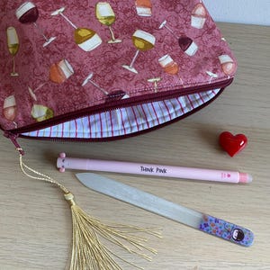 Può includere: Una pochette rosa con cerniera con una stampa di bicchieri di vino. La pochette è aperta e mostra una fodera a righe. La pochette è su una superficie di legno con una penna rosa, una lima per unghie, un fiocco dorato e un cuore rosso.