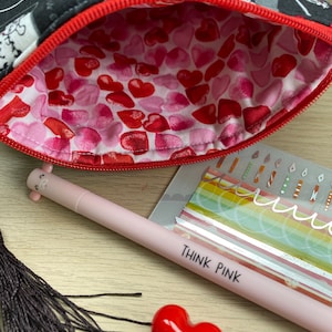 Pu&ograve; includere: Una pochette con cerniera nera e bianca con una cerniera rossa. L'interno della pochette &egrave; foderato con un motivo a cuori rosa e rossi. La pochette &egrave; aperta e una penna rosa con le parole "Think Pink" &egrave; appoggiata su una superficie di legno. Un oggetto rosso a forma di cuore e un fiocco nero sono anche sulla superficie.