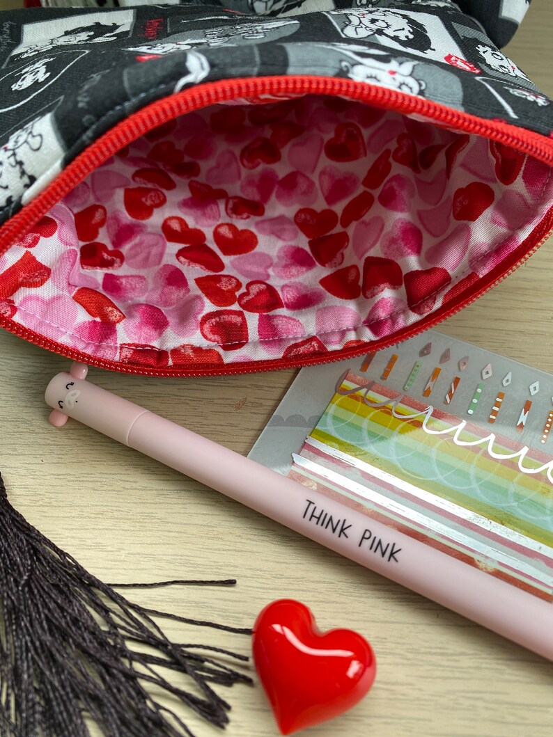 Pu&ograve; includere: Una pochette con cerniera nera e bianca con una cerniera rossa. L'interno della pochette &egrave; foderato con un motivo a cuori rosa e rossi. La pochette &egrave; aperta e una penna rosa con le parole "Think Pink" &egrave; appoggiata su una superficie di legno. Un oggetto rosso a forma di cuore e un fiocco nero sono anche sulla superficie.
