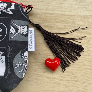 Pu&ograve; includere: Borsa in tessuto nero e bianco con chiusura a zip e un ciondolo a forma di cuore rosso. La borsa presenta un'immagine di cartone animato di Betty Boop e il testo "Made by F+F".