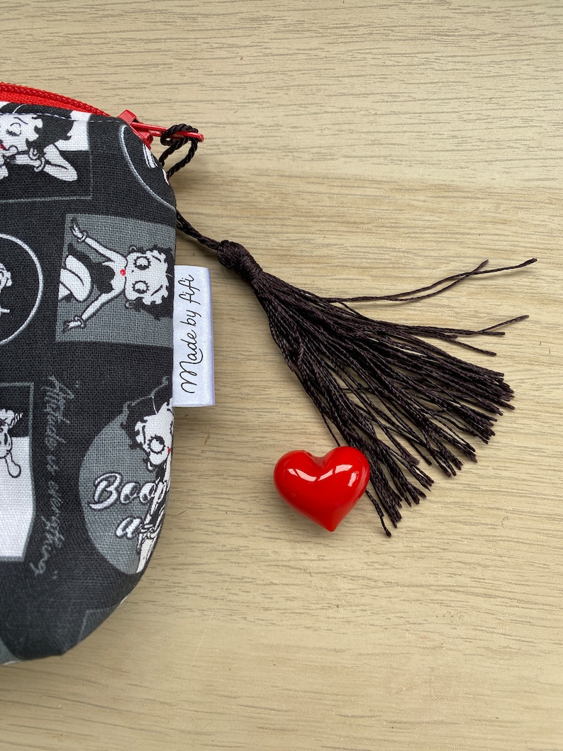 Pu&ograve; includere: Borsa in tessuto nero e bianco con chiusura a zip e un ciondolo a forma di cuore rosso. La borsa presenta un'immagine di cartone animato di Betty Boop e il testo "Made by F+F".