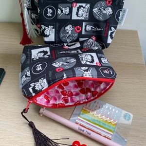 Pu&ograve; includere: Due pochette con cerniera nere e bianche con una stampa di un personaggio dei cartoni animati. Le pochette sono foderate con un tessuto rosso con cuori bianchi. Le pochette sono su una superficie di legno con una penna rosa, un fiocco nero e un cuore rosso.