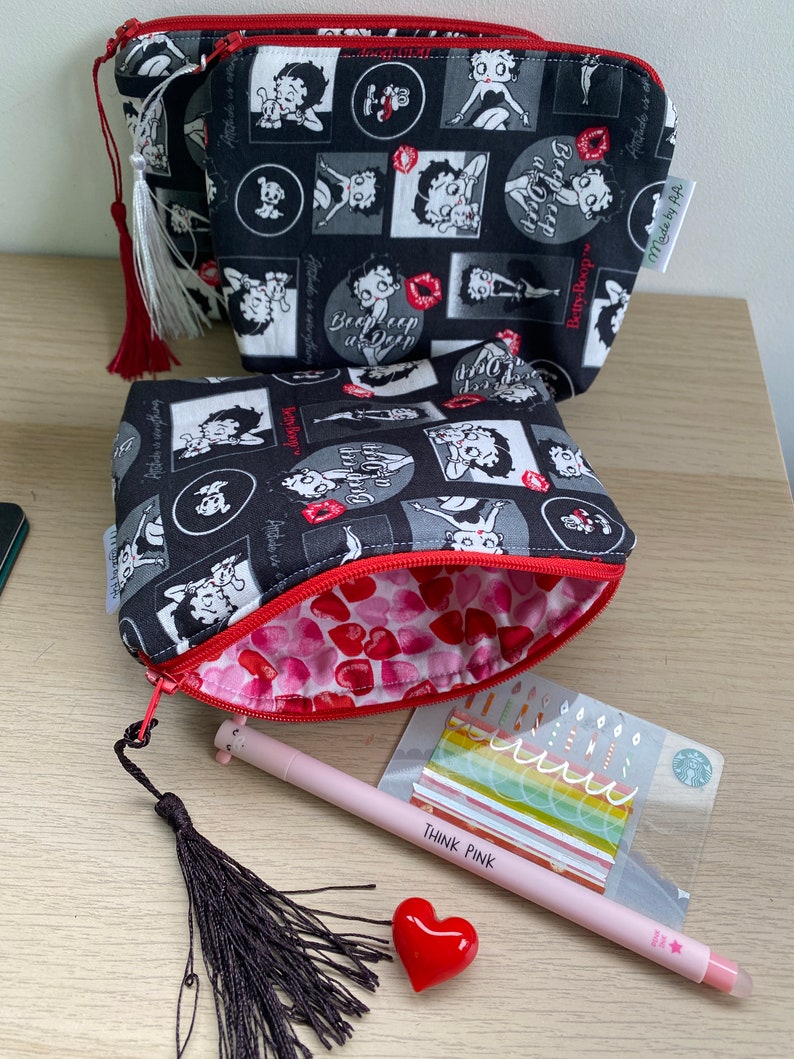 Pu&ograve; includere: Due pochette con cerniera nere e bianche con una stampa di un personaggio dei cartoni animati. Le pochette sono foderate con un tessuto rosso con cuori bianchi. Le pochette sono su una superficie di legno con una penna rosa, un fiocco nero e un cuore rosso.