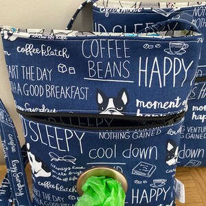 Può includere: Una borsa in tessuto blu e bianco con una chiusura a zip e un dispenser per i sacchetti per i rifiuti dei cani. La borsa ha una varietà di testo stampato su di essa, tra cui "coffee klatch", "coffee beans", "happy", "sleepy" e "cool down".