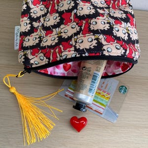 Pu&ograve; includere: Una pochette con cerniera nera e rossa con una stampa di cartoni animati di una donna con occhi grandi. La pochette &egrave; aperta e contiene un tubo di crema per le mani, un piccolo cuore rosso e un fiocco giallo. La pochette ha un'etichetta che dice "Made by P.A."