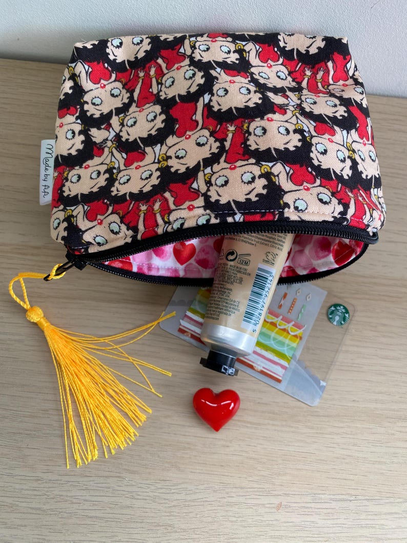 Pu&ograve; includere: Una pochette con cerniera nera e rossa con una stampa di cartoni animati di una donna con occhi grandi. La pochette &egrave; aperta e contiene un tubo di crema per le mani, un piccolo cuore rosso e un fiocco giallo. La pochette ha un'etichetta che dice "Made by P.A."