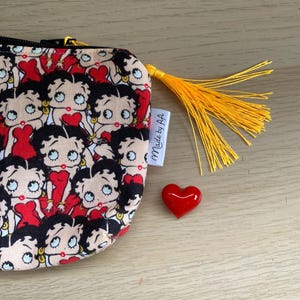 Pu&ograve; includere: Una piccola pochette beige con cerniera e un fiocco giallo. La pochette &egrave; ricoperta da un motivo ripetitivo di personaggi dei cartoni animati di Betty Boop. La pochette ha un'etichetta bianca che dice "Made by A+A". Un cuore rosso &egrave; seduto accanto alla pochette.