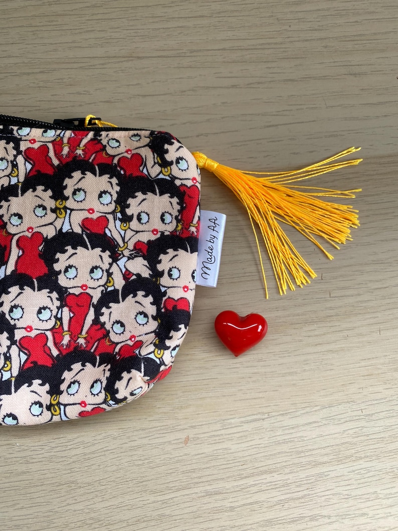 Pu&ograve; includere: Una piccola pochette beige con cerniera e un fiocco giallo. La pochette &egrave; ricoperta da un motivo ripetitivo di personaggi dei cartoni animati di Betty Boop. La pochette ha un'etichetta bianca che dice "Made by A+A". Un cuore rosso &egrave; seduto accanto alla pochette.