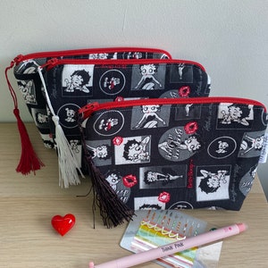 Pu&ograve; includere: Tre pochette con cerniera nera e bianca con una cerniera rossa e un fiocco rosso. Le pochette presentano un motivo ripetuto di immagini di Betty Boop. Le pochette sono su una superficie di legno con una penna rosa e un oggetto a forma di cuore.