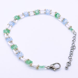 Op de afbeelding: Een delicate kralenarmband met afwisselende clusters van lichtblauwe en groene bloemvormige kralen. De armband is geregen op een wit koord en heeft een zilverkleurige sluiting en verlengketting. De lengte van de armband is verstelbaar.
