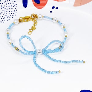 以下が含まれることがあります： パールと金のアクセントが付いた薄い青色のビーズブレスレット。ブレスレットは青いビーズで作ったリボンが付いている。