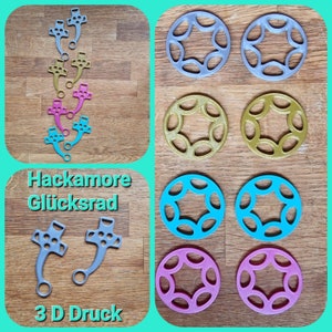 Może przedstawiać: Zestaw 3D-wydrukowanych części do Hackamore Glückrad, rodzaju koła obrotowego. Części są w różnych kolorach, w tym srebrnym, złotym, różowym, niebieskim i turkusowym. Części są ułożone na drewnianej powierzchni.