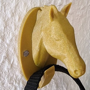 Op de afbeelding: Een gouden paardenkop wandhaak met een zwarte band die eraan hangt.
