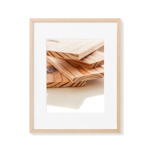 May include: A framed photograph showing a stack of unfinished wooden planks. The planks are a warm, natural wood tone with visible grain and knots. The frame is a light-coloured wood, and the background is white.