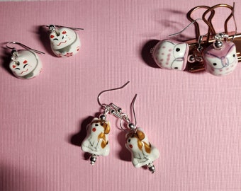 May include: Three pairs of earrings with ceramic charms. The first pair features a white cat charm with pink and gold accents. The second pair features a white owl charm with pink and black accents. The third pair features a white dog charm with brown accents.