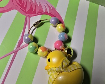 May include: A keychain featuring a yellow duck figurine with a yellow and white checkered ribbon tied around its neck. The duck is attached to a circular wire ring adorned with several multicolored, pastel-toned beads and a small gold-colored bell.