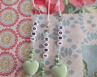 May include: Three beaded phone charms with the name "HOBI" spelled out in white letter beads. Each charm has a light green heart-shaped bead and is strung on a white cord. The background features a floral pattern.