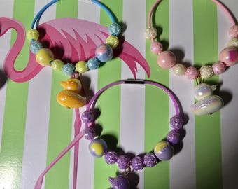 May include: Three circular necklaces featuring colorful plastic beads and a central rubber duck charm. One necklace has a blue band with yellow and blue beads, one has a pink band with pink and white beads, and one has a purple band with purple beads.