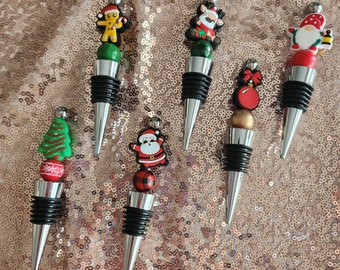 May include: Six different Christmas themed bottle stoppers. The stoppers are silver with a black top and feature various Christmas characters and designs. The characters include a gingerbread man, a reindeer, a snowman, a Santa Claus, a Christmas tree, and a Christmas ornament.