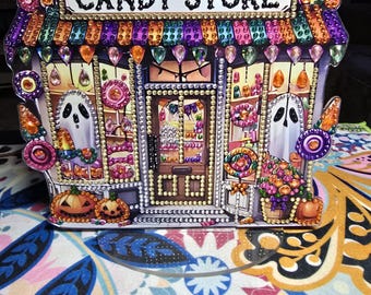 May include: A 3D diamond painting of a haunted candy store with a sign that reads "Haunted Candy Store". The store is decorated with ghosts, pumpkins, and candy. The store is lit up with string lights.