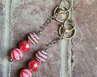 May include: A red and white candy cane themed keychain with silver key rings. The keychain features round beads with red and white stripes and red and white glitter.