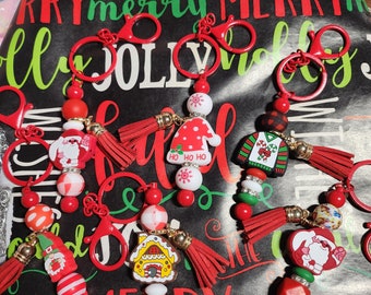 May include: A set of five Christmas-themed keychains with red, white, and green beads, red tassels, and charms featuring a gingerbread house, a snowman, a Santa Claus, a reindeer, and a Christmas sweater. The keychains are displayed on a black background with the words "Merry" and "Jolly" printed in red and green.